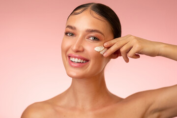 Hydration and skin care concept. Beauty portrait of young beautiful woman applying cream on her cheek and smiling, posing over pink background