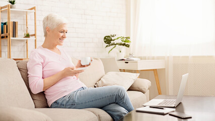 Modern morning. Positive senior woman enjoying coffee and watching news on laptop, sitting on couch at home, free space