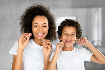 A cheerful African American mother and her young son are standing together in a well-lit, contemporary bathroom, wearing white t-shirts while they both floss their teeth