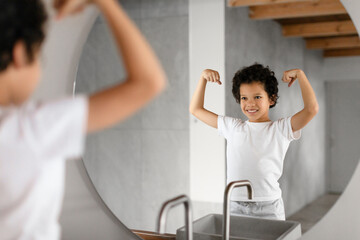 African American young child is standing in front of a bathroom mirror, proudly flexing their muscles and exhibiting a joyful smile. The bathroom has a modern design with visible wooden beams