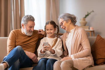 A family consisting of a mother, father, and child are seated on a couch indoors. They are engrossed in looking at a cell phone screen together, showing a close bond and shared interests