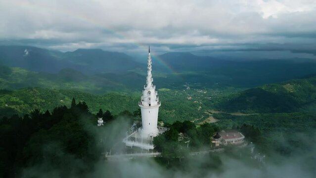 Drone flies over Ambuluwawa Tower, revealing rainbow in misty Sri Lanka mountains. Scenic travel destination panorama, vibrant green landscapes under cloudy skies, offering breathtaking views. Aerial