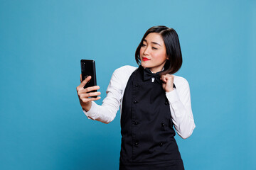 Attractive asian waitress adjusting bow tie and looking at smartphone front camera while taking...