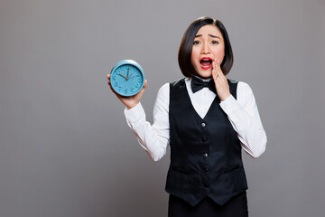 Anxious asian waitress panicking while holding circle dial alarm clock and looking at camera with open mouth. Young terrified woman receptionist showing timer watch portrait