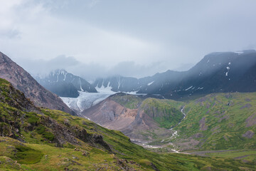 Dramatic misty view to green hills and rocks against big glacier tongue and large mountain range...