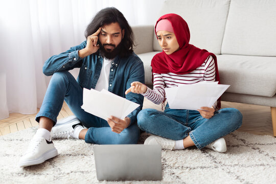 A young indian man with a beard and a woman wearing a hijab sit closely on the floor with papers in hand, looking worried as they review financial documents together beside an open laptop