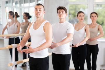 Group of male and female dancers stand in first ballet pose at barre in dance class