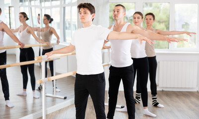 Group of male and female dancers stand in second ballet pose at barre in dance class