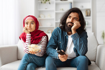 Unhappy indian young couple is lounging on a cozy sofa in a well-lit living room, seemingly...