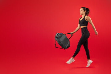 A young woman dressed in black athletic wear confidently takes a stride, holding a sports bag in...