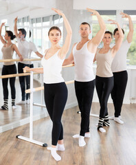 Group of male and female dancers stand in fourth ballet pose at barre in dance class
