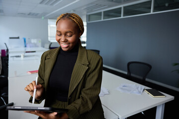 Young businesswoman in businesswear smiling while using digital tablet and digitized pen on desk in office