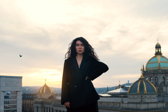 Attractive young curly brunette woman in a black jacket without lingerie sits on a rooftop against a historic building during a cozy sunset