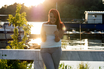 Teenager with red hair relaxing near the river. Beautiful long haired redhead is using her digital tablet near the river bank during sunset