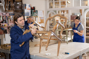 Middle-aged man restorer applies primer to surface of wooden vintage chair to repair damage,...