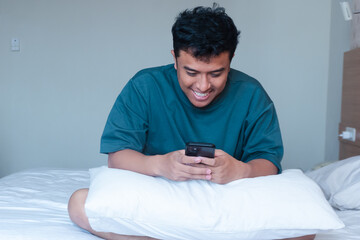 Smiling adult Asian man replying to chat on cellphone while sitting on bed