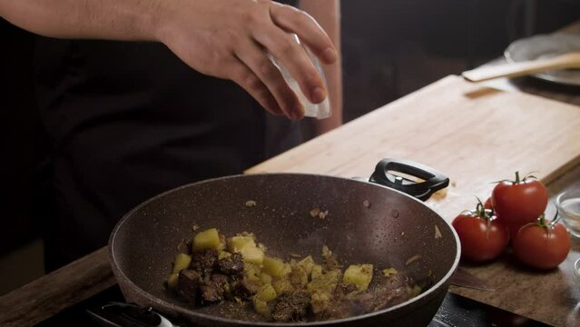 Enjoy the skill of the chef as he sprinkles seasonings on a delicious dish of meat and potatoes, showcasing his culinary talent. Observe as the ingredients are carefully combined and brought to