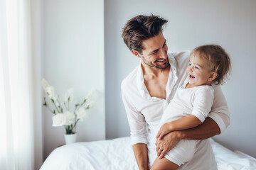 Fototapeta premium Lifestyle father holding his laughing baby stands near the window in white bedroom. 