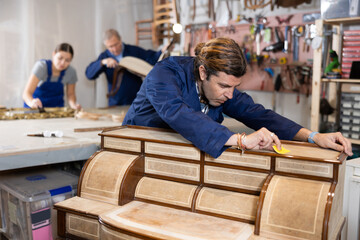In restoration showroom, middle-aged man carefully polishes tabletop of old chest of drawers with...