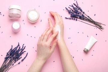 Woman applying hand cream and lavender flowers on pink background, top view