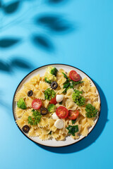 Delicious pasta salad with farfalle, cherry tomatoes, olives, mozzarella, broccoli, and parsley on a blue background.