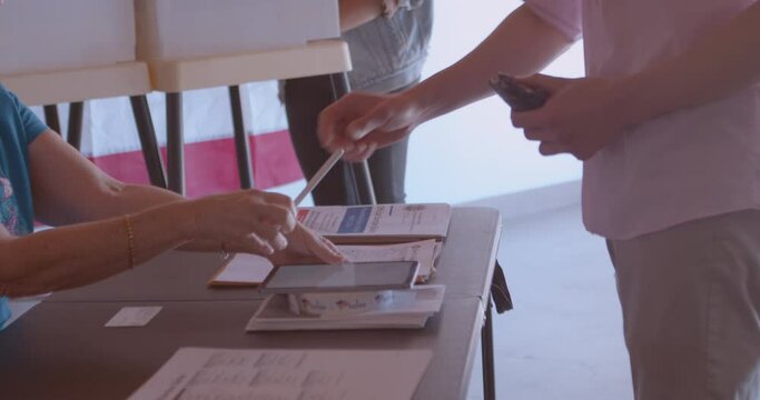 A mature female poll worker processing a male voter with a tablet at a polling station draped with American flags.