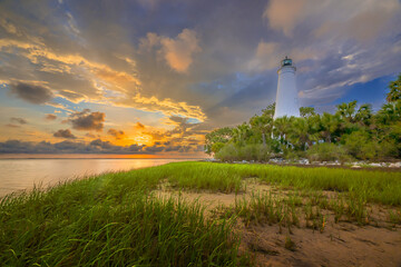 "As the sun sets behind St. Marks Lighthouse, casting a warm glow over the grasses and palm trees, the cloudy sky paints a mesmerizing backdrop, creating a picturesque scene of coastal beauty."