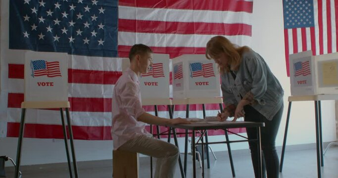 Wide shot of a male poll worker processing voters at a polling station draped with American flags. 