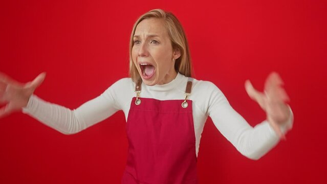 Enraged young woman, blonde, standing over red background, screaming in anger; wearing apron; furious, frustrated, aggressive