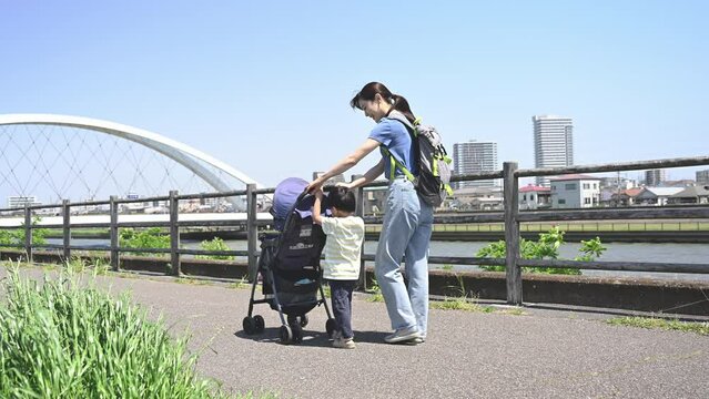 晴天の川沿いの公園でベビーカーで散歩するママと兄弟の親子(3人家族)の全身　春夏のお出かけやママの通勤などのイメージスロー動画　後ろ姿の顔無し