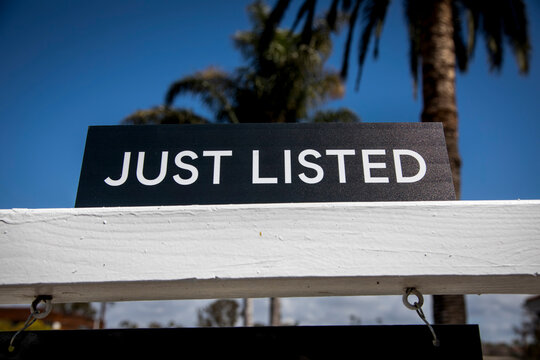 A black realtor sign with white letters advertising a new home listing  Plam trees are in the background