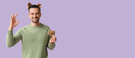 Handsome man in reindeer horns with tasty cookie showing OK on lilac background