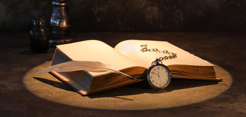 Open old book with clock, feather and burning candle on table against dark background