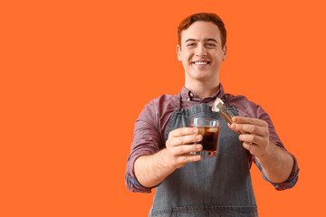 Young male bartender putting ice into glass with cocktail on orange background