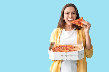Young woman eating piece of tasty pizza and holding cardboard box on blue background