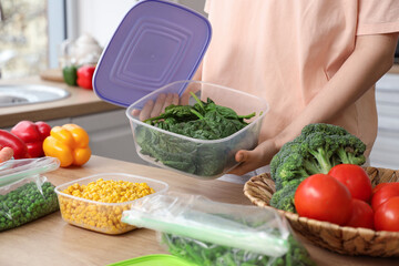 Woman holding plastic container with spinach for freezing at table in kitchen