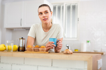 Dreamy young woman standing in home kitchen with cup of coffee