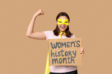 Young woman in superhero costume holding cardboard with text WOMEN'S HISTORY MONTH on beige...