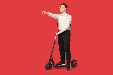 Young man with modern electric kick scooter pointing at something on red background
