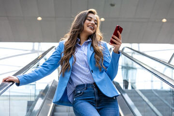 Smiling young business woman wearing suit standing on urban escalator using applications on cell...