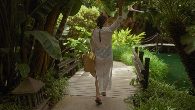 A tranquil scene of a young woman walking on a wooden bridge amidst lush greenery in a tropical balinese resort.