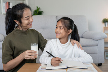 Asian young little girl learn at home. Do homework with kind mother help, encourage for exam. Mom pass on a glass of milk to daughter. Girl happy Homeschool. Mom teach and advice education together.