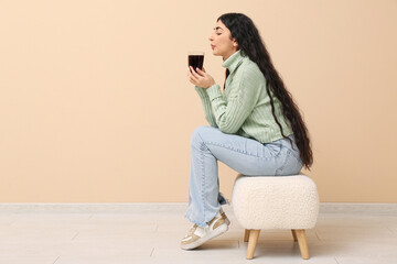 Beautiful young woman with glass cup of hot tea sitting on pouf near beige wall