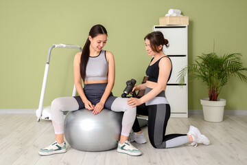 Female physiotherapist massaging young woman's thigh with percussive massager on fitball in...