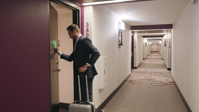 A businessman in a tailored gray suit is captured from the side as he uses smartphone as a key card to unlock his hotel room door, suitcase by his side, portraying a scene of convenience and security 