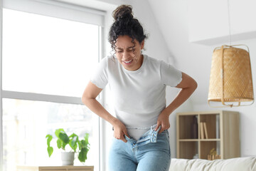 Young African-American woman trying to put on tight jeans at home. Diet concept