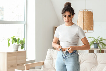 Upset African-American woman trying to button tight jeans at home. Diet concept