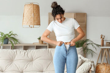 Young African-American woman trying to button tight pants at home. Diet concept