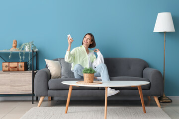 Smiling young woman with air conditioner remote control sitting on sofa near blue wall