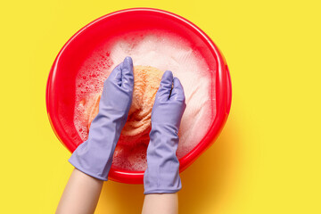 Woman in rubber gloves washing clothes in plastic basin on yellow background